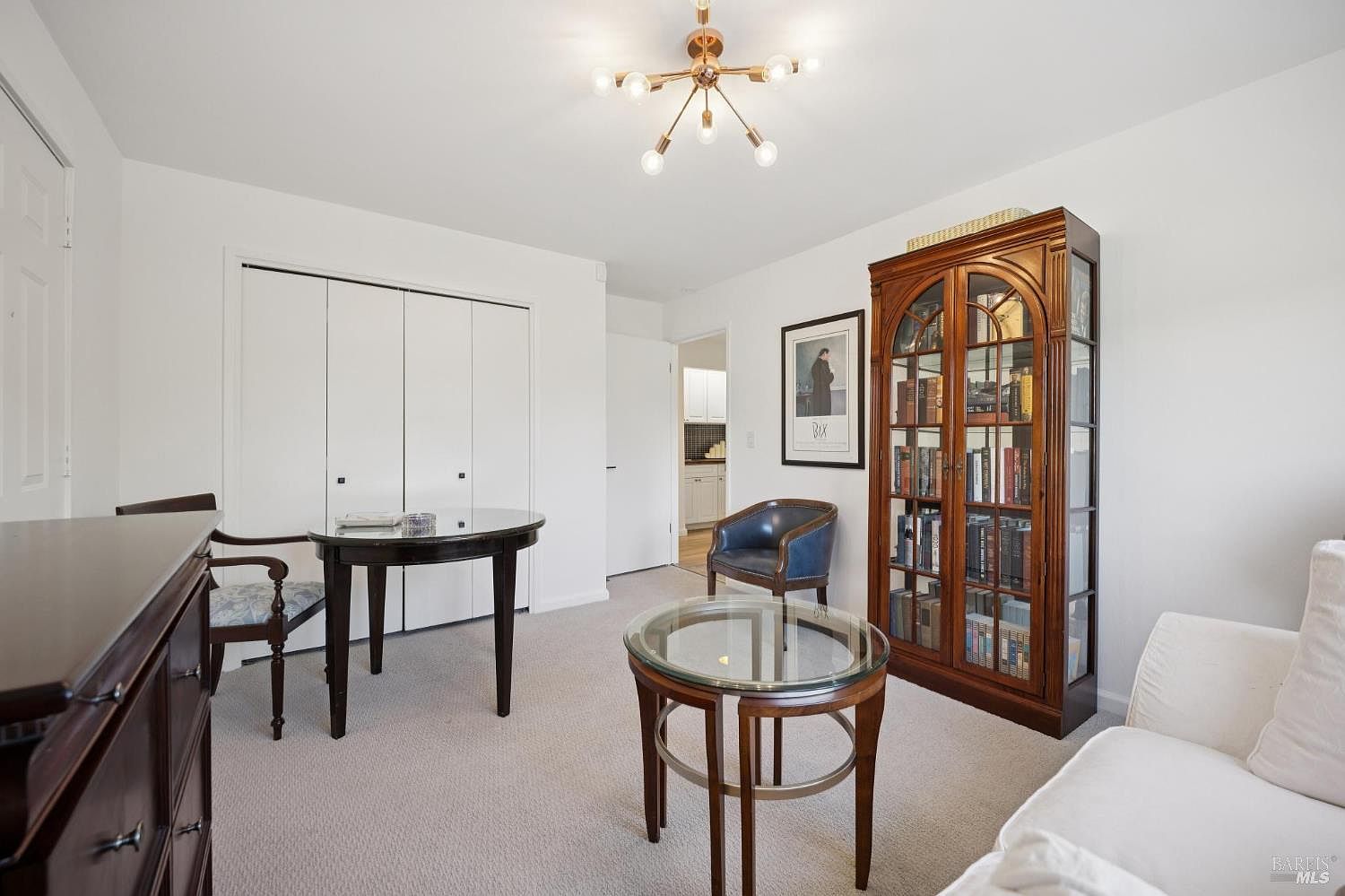 This bright and versatile room functions as a home office or sitting area, featuring neutral carpeting and crisp white walls. The space is furnished with a round glass-topped table, a classic wooden bookcase, and a blue accent chair, all illuminated by a modern, multi-arm chandelier. A doorway in the background provides a glimpse into an adjacent kitchen, enhancing the open and connected feel of the home.