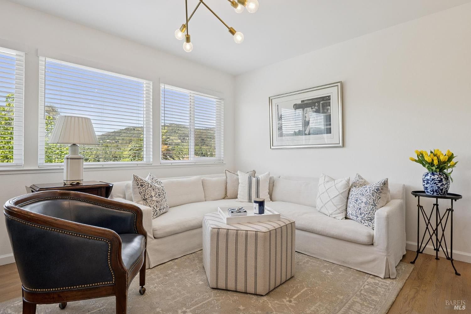This bright and inviting living room features a large, cream-colored sectional sofa arranged around a striped, hexagonal ottoman. A dark leather armchair sits to the left, while a small side table with a vase of yellow tulips adds a pop of color to the right. Large windows with white blinds allow for ample natural light and provide a scenic view of the surrounding hills.