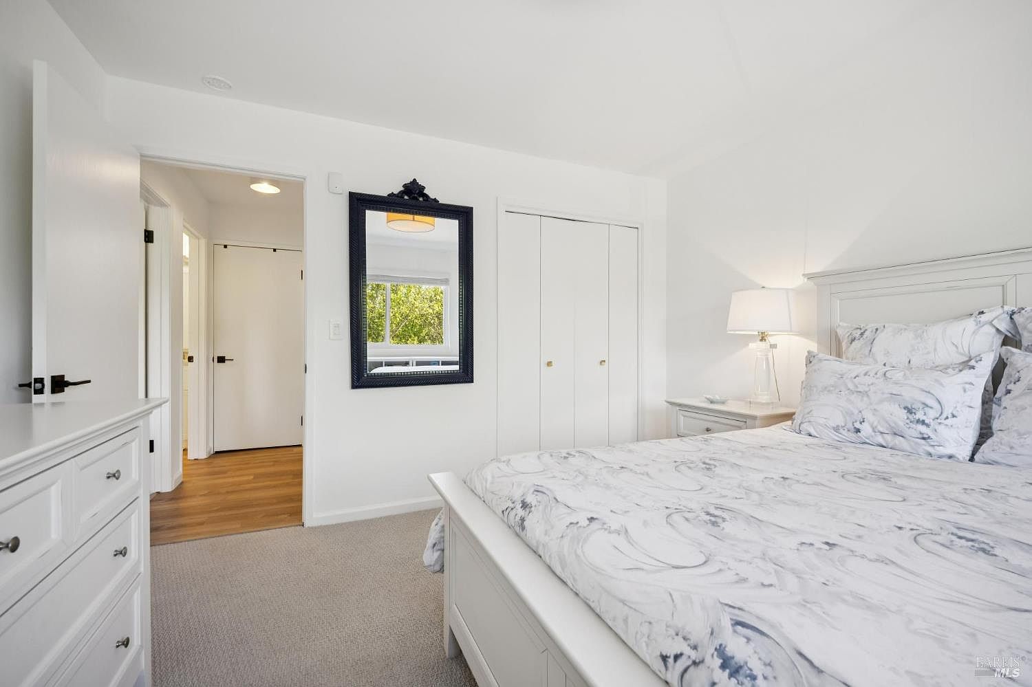 This bright and airy bedroom features a white bed frame with patterned bedding and a matching white dresser. A dark-framed mirror hangs on the wall, reflecting a view of the outdoors, while a bedside lamp provides warm lighting. The room is finished with neutral carpeting and leads into a hallway with light wood flooring.