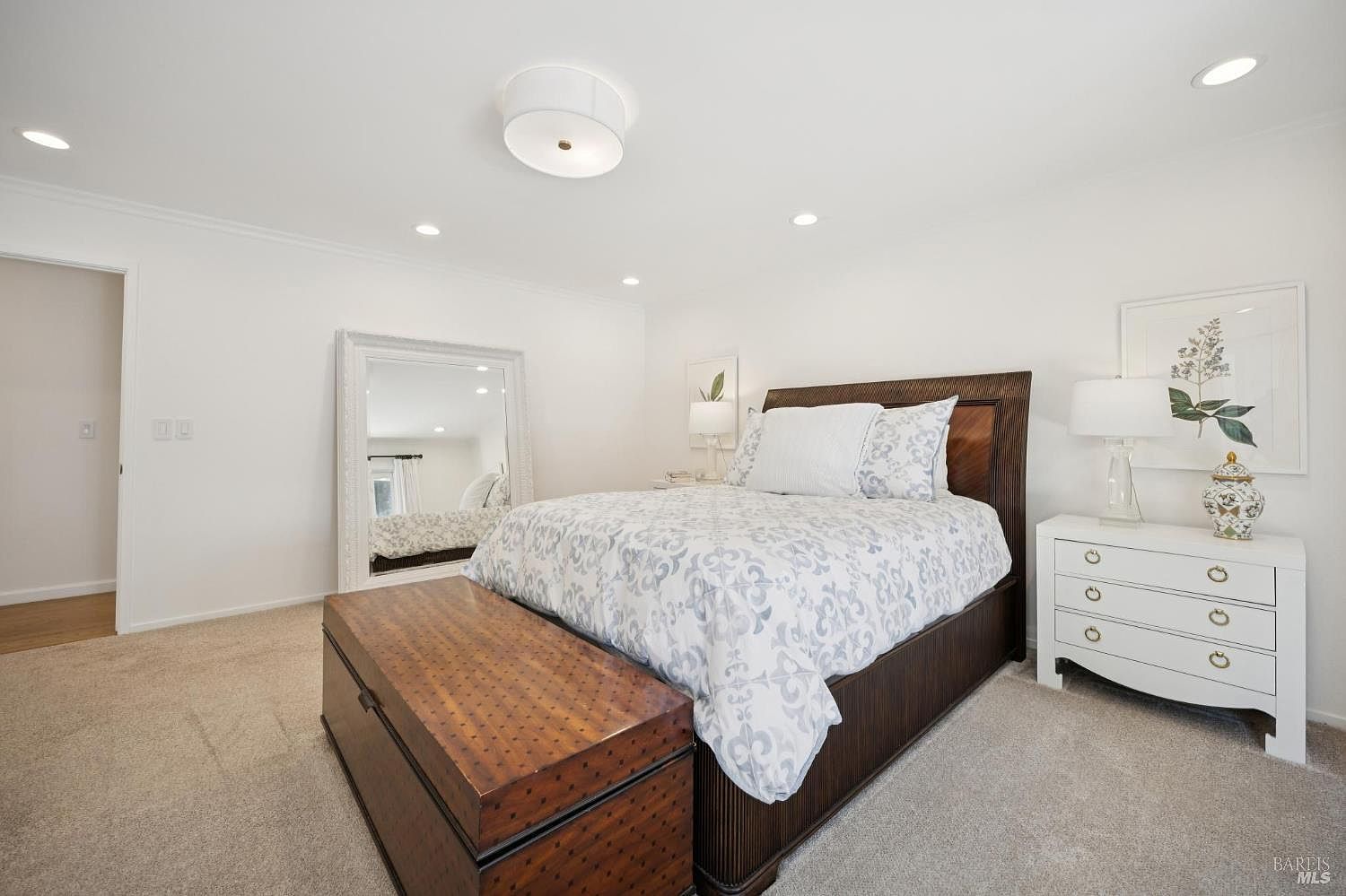 This primary bedroom features a dark wood bed frame with a patterned duvet and a matching wooden storage chest at the foot of the bed. A white nightstand with a lamp and decorative vase sits to the right, while a large floor mirror leans against the wall on the left. The room is finished with neutral carpeting, white walls, and recessed lighting, creating a bright and serene atmosphere.
