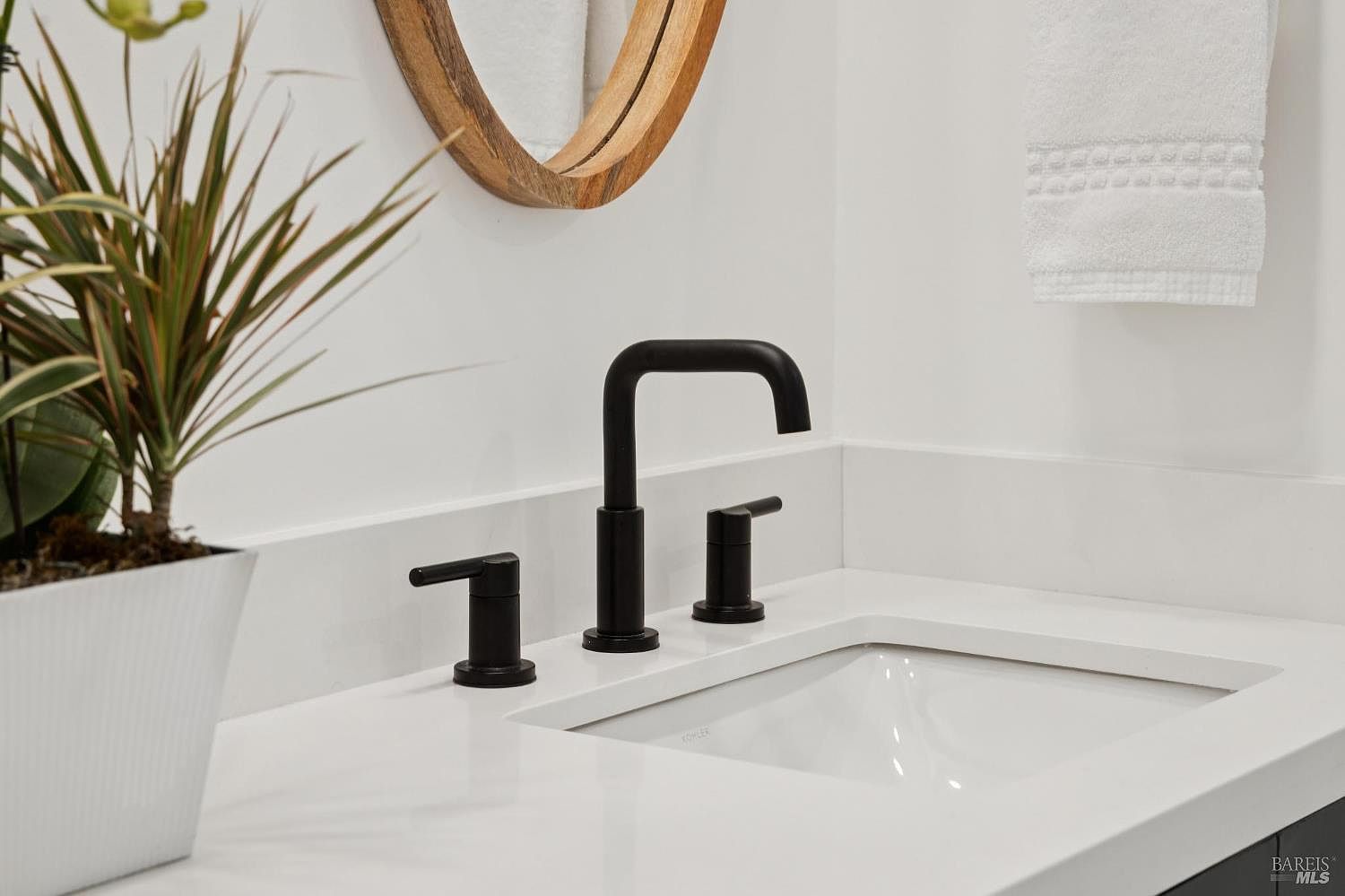 This close-up shot highlights a modern bathroom vanity featuring a sleek, matte black widespread faucet set against a clean white countertop. A portion of a round, light-wood framed mirror is visible above, reflecting the bright, minimalist space, while a white towel hangs on the wall to the right. A potted plant adds a touch of greenery to the left, contributing to the fresh and contemporary aesthetic of the room.