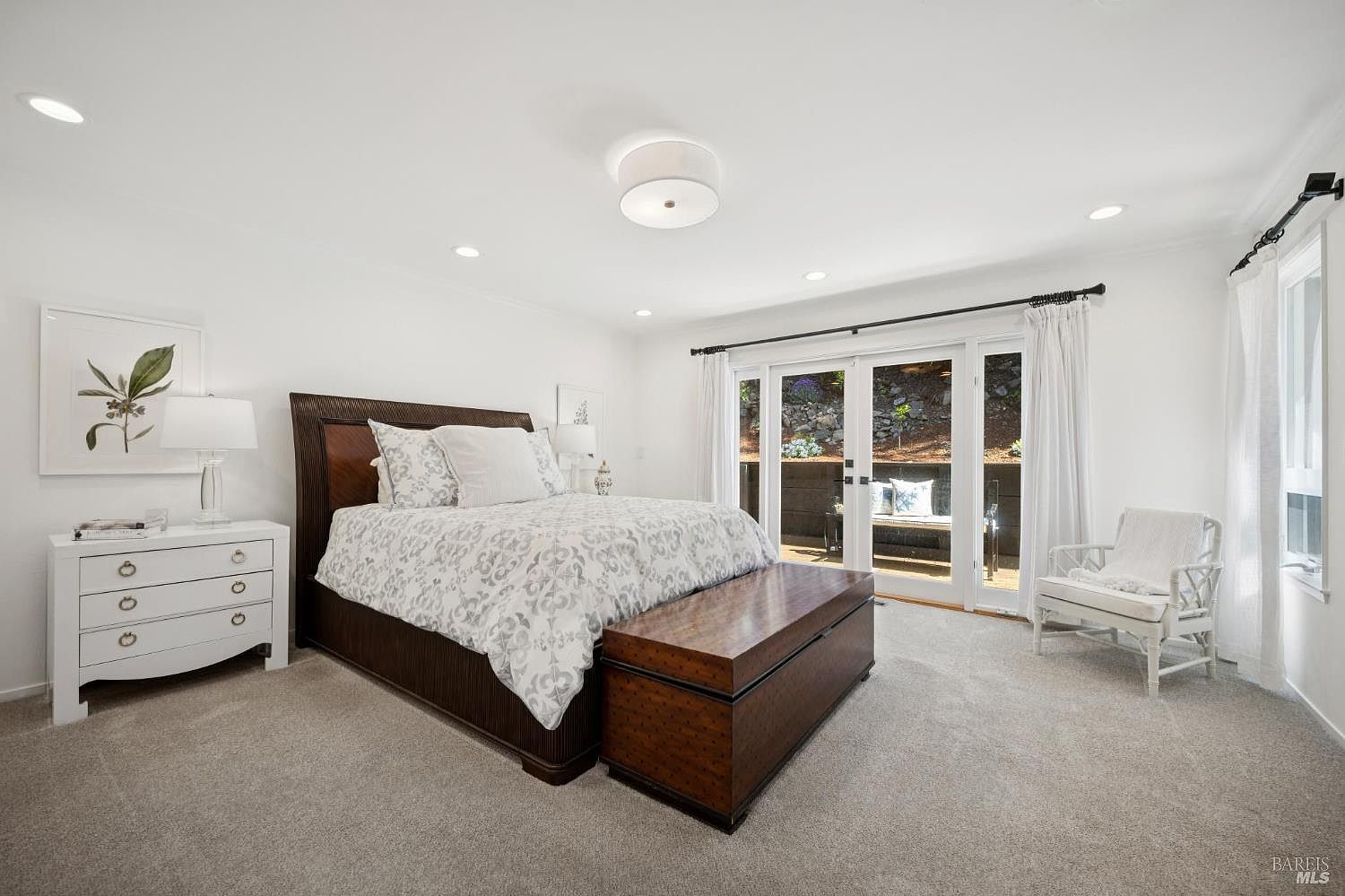 This primary bedroom features a large, dark wood-framed bed as the centerpiece, complemented by a matching storage chest at the foot of the bed. A white dresser with a lamp and framed artwork sits to the left, while a white wicker chair is positioned near the glass doors that lead to an outdoor patio. The room is bright and airy, with neutral carpeting and white walls, creating a serene and inviting atmosphere.