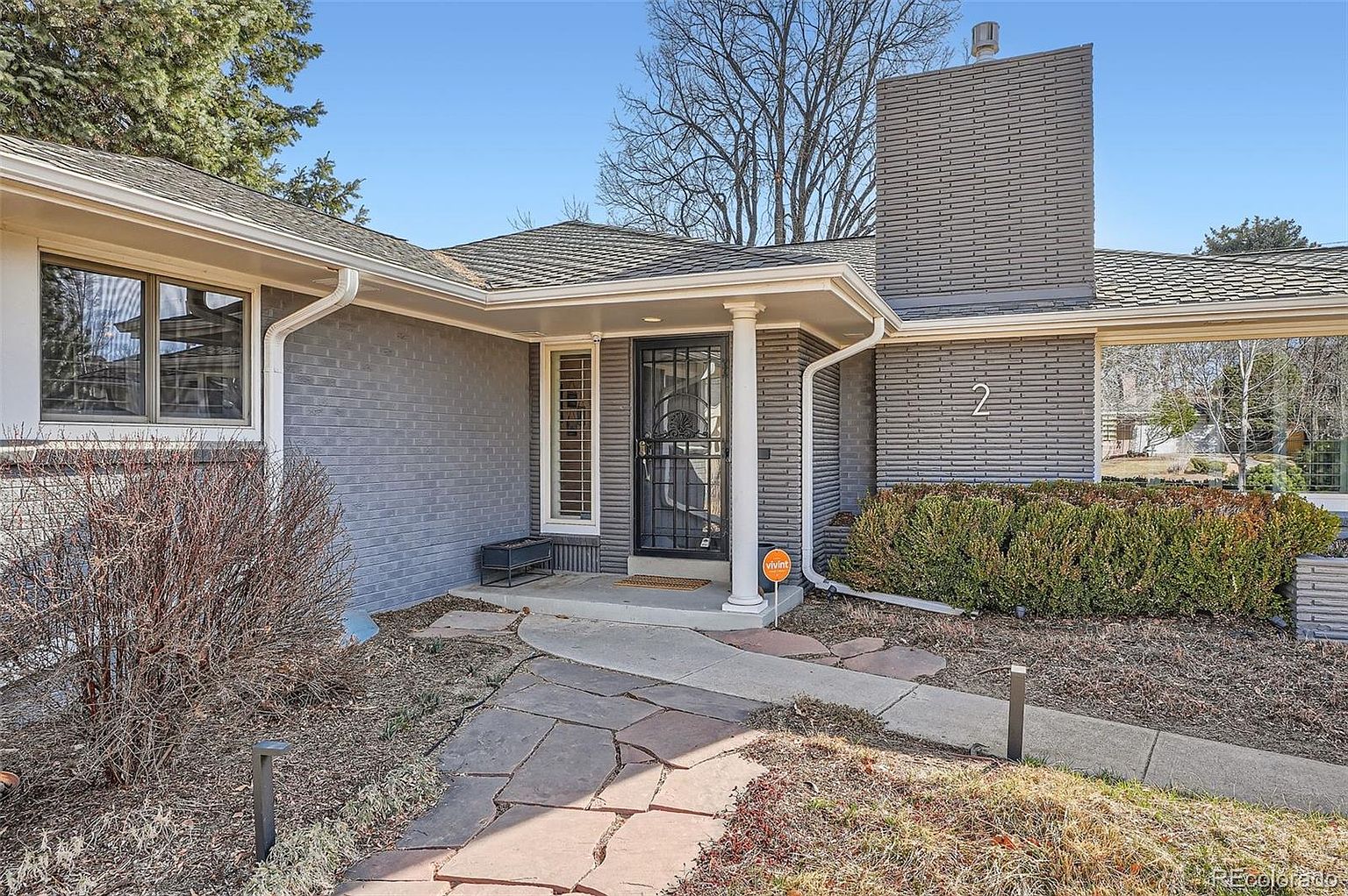 The image showcases the entryway of a single-story home, featuring a gray brick facade and a prominent chimney. A black security door is framed by white pillars, leading to a stone pathway. The landscaping includes neatly trimmed bushes and decorative lighting, creating a welcoming and well-maintained appearance.