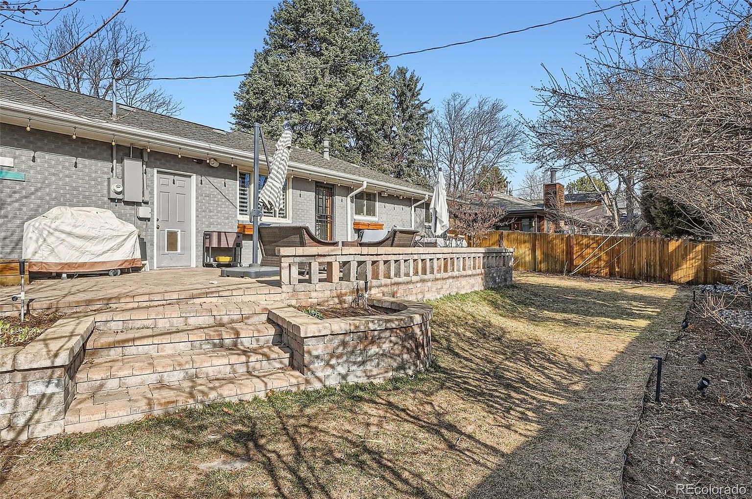 This image showcases a well-maintained backyard featuring a brick patio with multiple levels and a low brick wall. The yard has a mix of grass and garden areas, with mature trees providing shade. A wooden fence encloses the property, offering privacy.