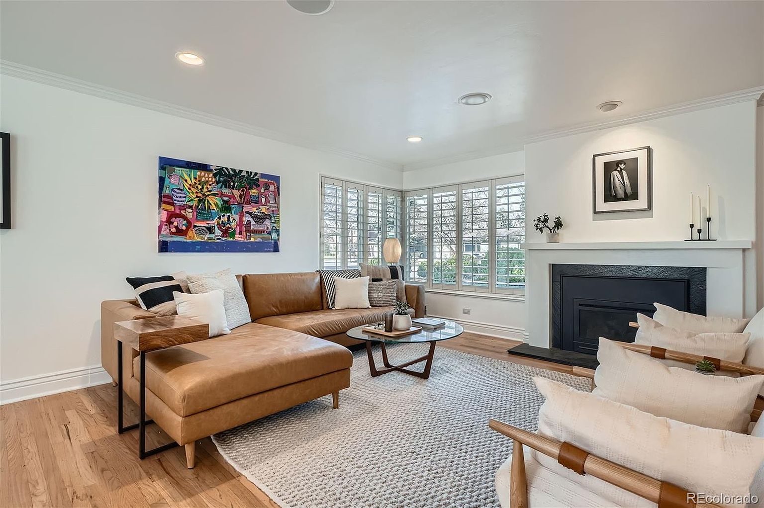 This is an interior shot of a living room featuring a brown leather sectional sofa, a round glass coffee table, and two armchairs with light-colored cushions. A fireplace with a black surround and white mantel is visible, along with a colorful painting on the wall. The room is well-lit with natural light from the windows and recessed lighting, creating a warm and inviting atmosphere.