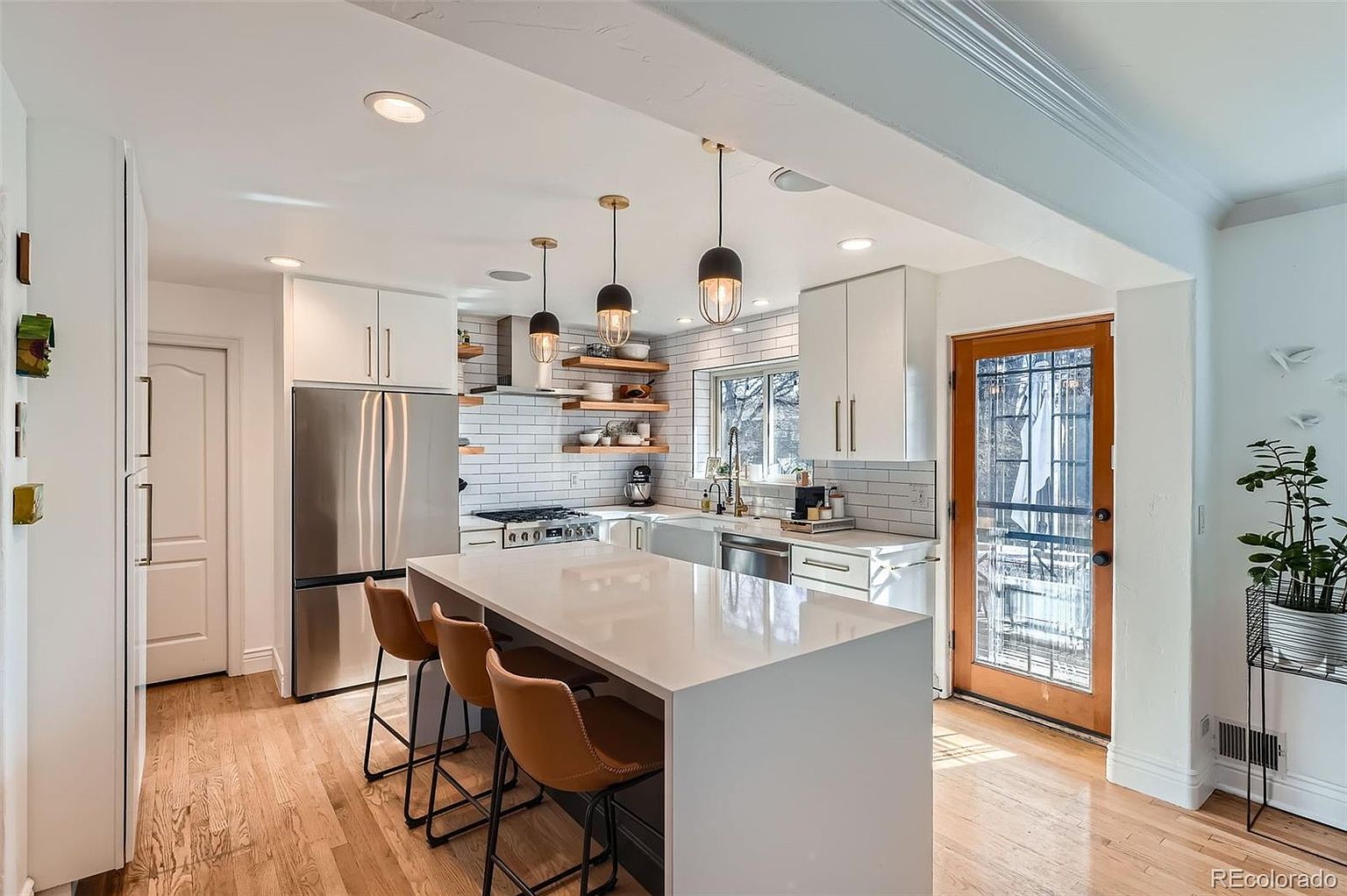 This is a bright and modern kitchen featuring white cabinetry, stainless steel appliances, and a large white island with seating. The kitchen is well-lit with pendant lighting and recessed lighting, and it has a window above the sink that provides natural light. The hardwood floors add warmth to the space, and the overall impression is clean and inviting.