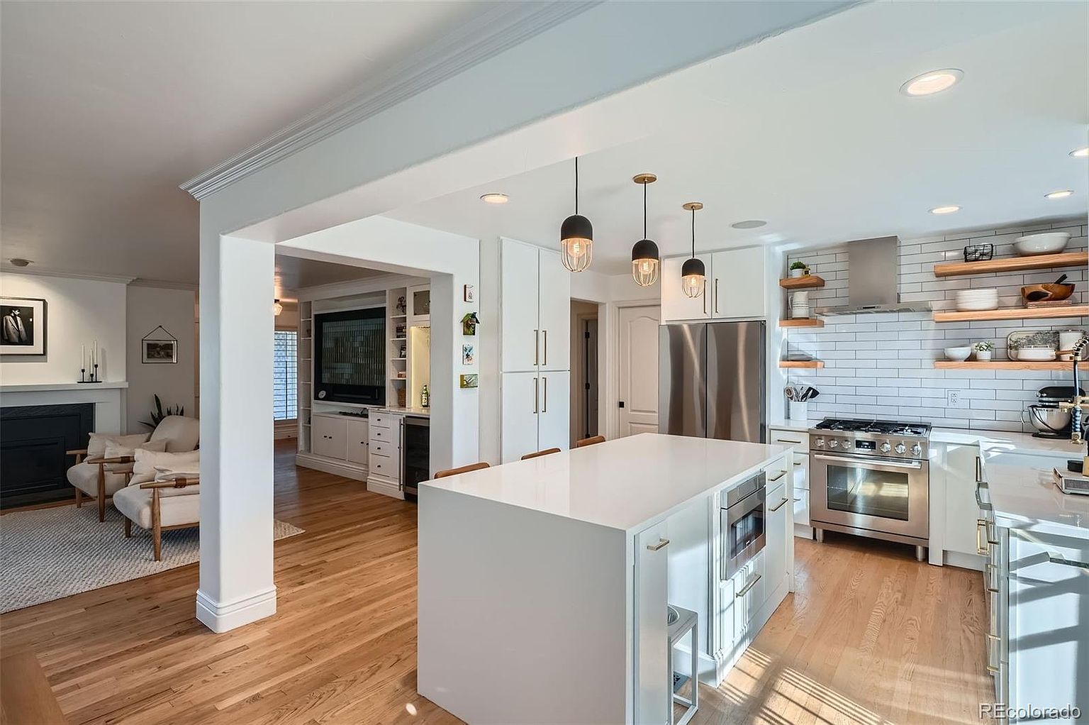 This is a bright and open kitchen featuring a large white island with built-in appliances, stainless steel appliances, and white subway tile backsplash with open wooden shelving. The kitchen seamlessly connects to a living area, creating an inviting and spacious atmosphere. The hardwood floors add warmth to the modern design.