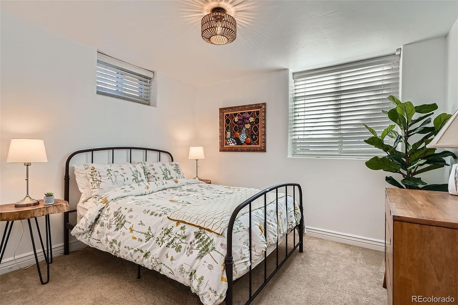 This is a cozy bedroom featuring a black metal bed frame with floral bedding, flanked by a wooden side table with a lamp and a small plant. A framed painting hangs on the wall above the bed, and natural light filters through the window with blinds. The room has a warm and inviting atmosphere, perfect for a guest room or a small bedroom.