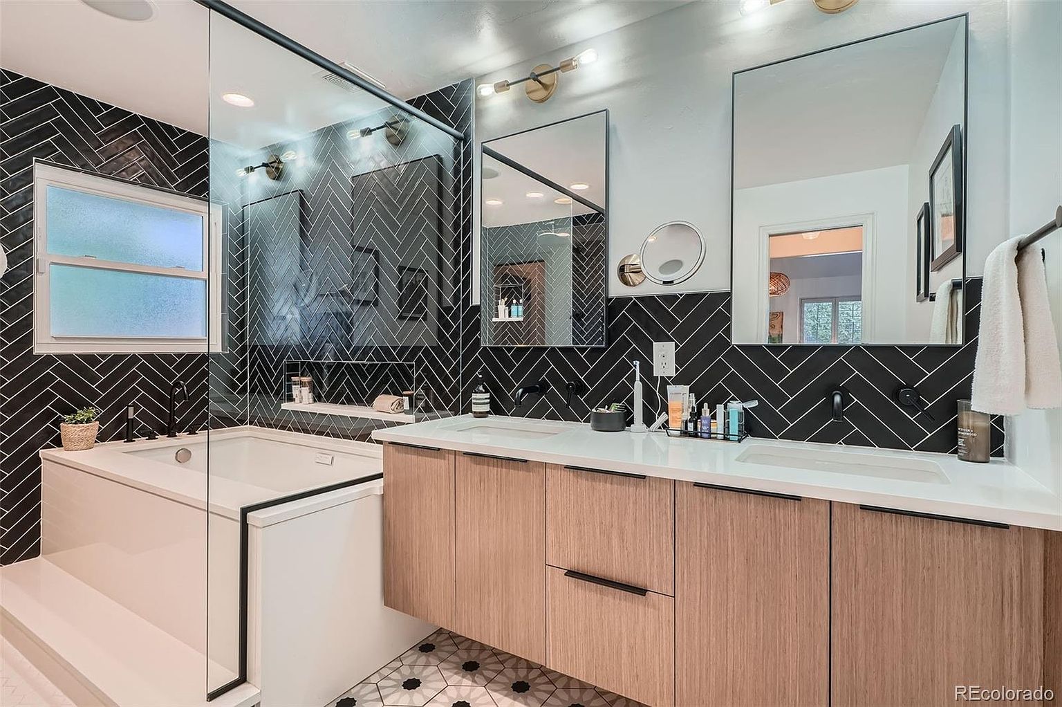 This is a modern primary bathroom featuring a double vanity with light wood cabinetry and a white countertop. The backsplash is black tile laid in a herringbone pattern, complemented by matching tile in the shower and around the bathtub. Two rectangular mirrors hang above the sinks, and the flooring is a patterned tile. The perspective is from the doorway, showcasing the entire vanity area and part of the tub.