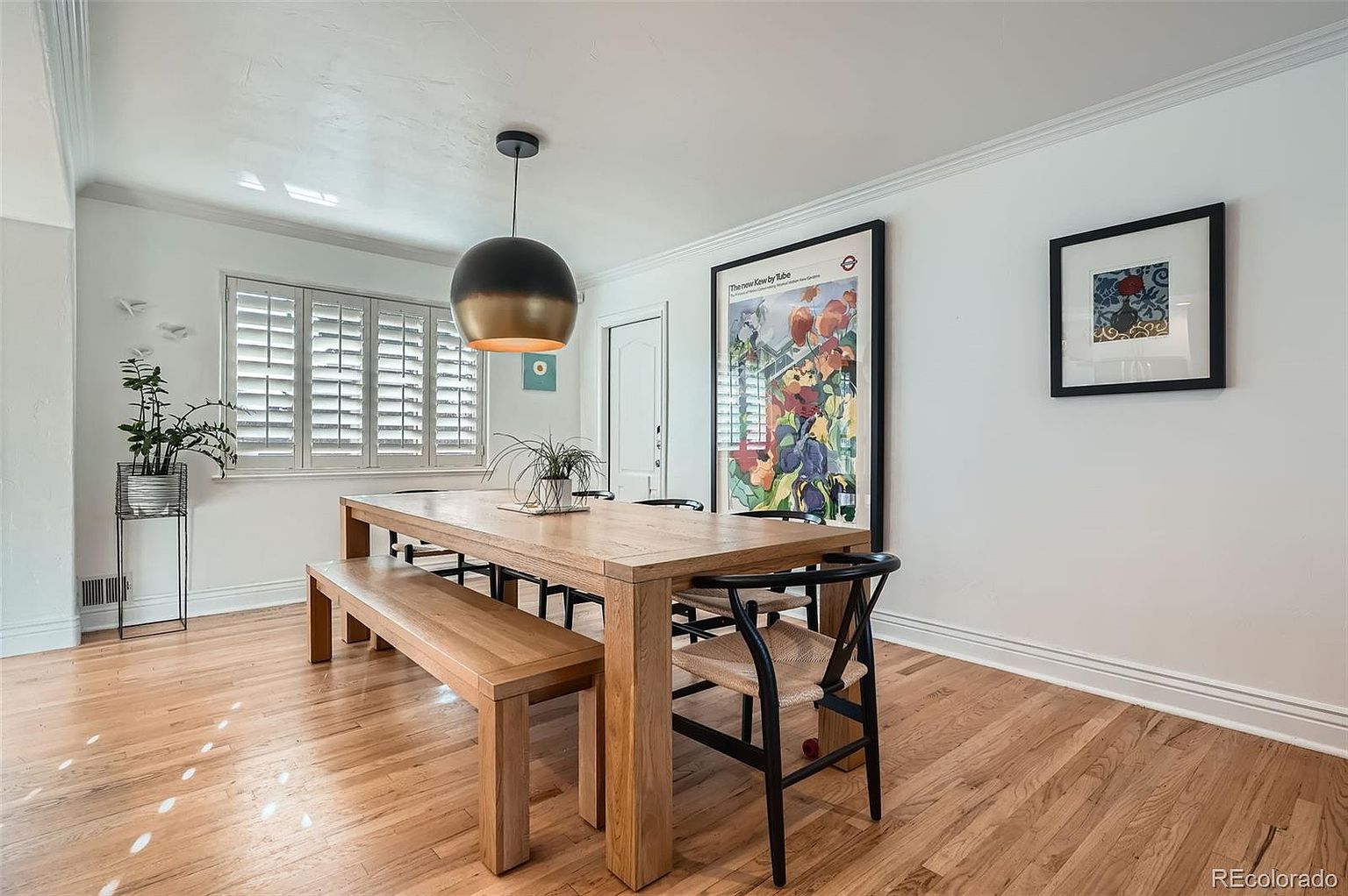 This is an interior shot of a dining room featuring a large wooden table with a bench and chairs. A modern pendant light hangs above the table, and artwork adorns the white walls. The room has hardwood floors and plantation shutters on the windows, creating a bright and inviting atmosphere.