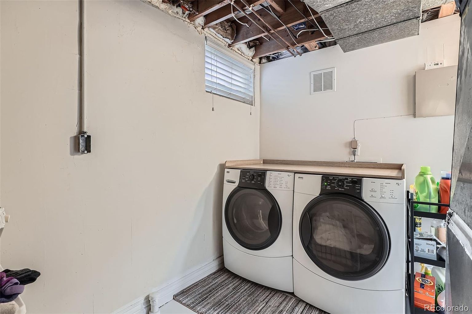 This laundry room features a front-loading washer and dryer set with a countertop above, providing a functional workspace. The room has neutral-colored walls and a small window for natural light. A storage rack with laundry supplies is visible on the right, adding to the room's utility.