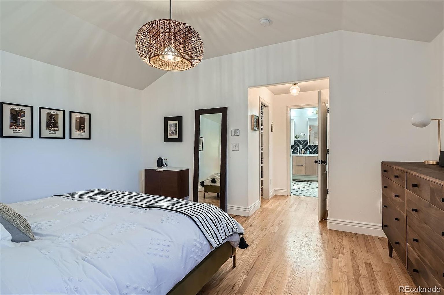 This is a bright and airy primary bedroom featuring hardwood floors, white walls, and a vaulted ceiling. The room is furnished with a bed dressed in white linens with a striped throw, a wooden dresser, and a full-length mirror. A unique woven pendant light hangs from the ceiling, adding a touch of modern style.