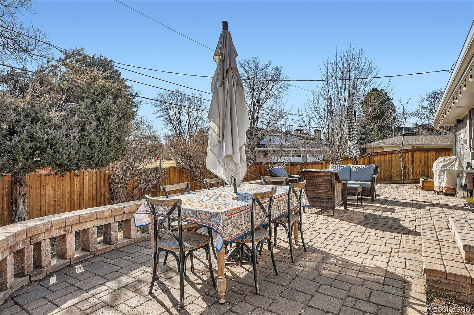 This image showcases a charming outdoor patio area, perfect for entertaining or relaxing. The patio is paved with brick and features a dining table with chairs, covered by a patterned tablecloth and shaded by a large, closed umbrella. Comfortable outdoor seating is arranged in the background, creating an inviting and functional outdoor living space.