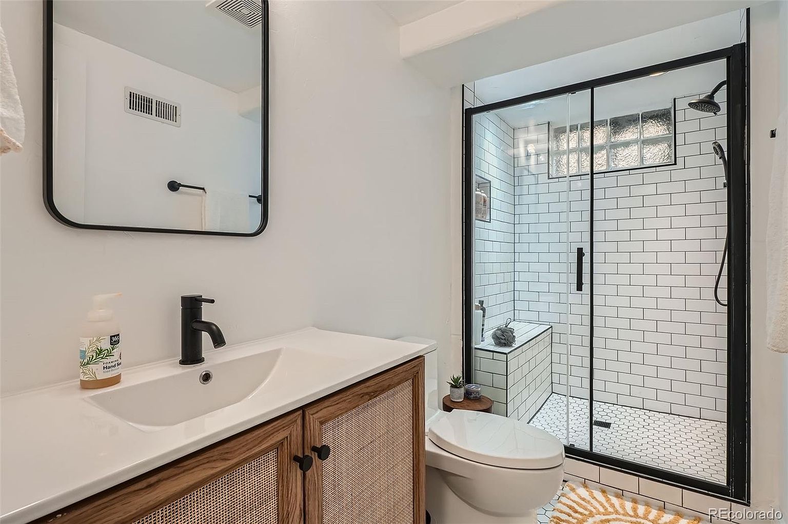 This is a well-lit bathroom featuring a modern design with a white vanity topped with a white countertop and a black faucet. The vanity has a wood frame with woven cane door panels. A black-framed mirror hangs above the vanity. The shower is enclosed with a black frame and glass, showcasing white subway tiles with dark grout and a hexagonal tile floor. A white toilet sits next to the shower, and a small wooden stool with a plant adds a touch of nature.