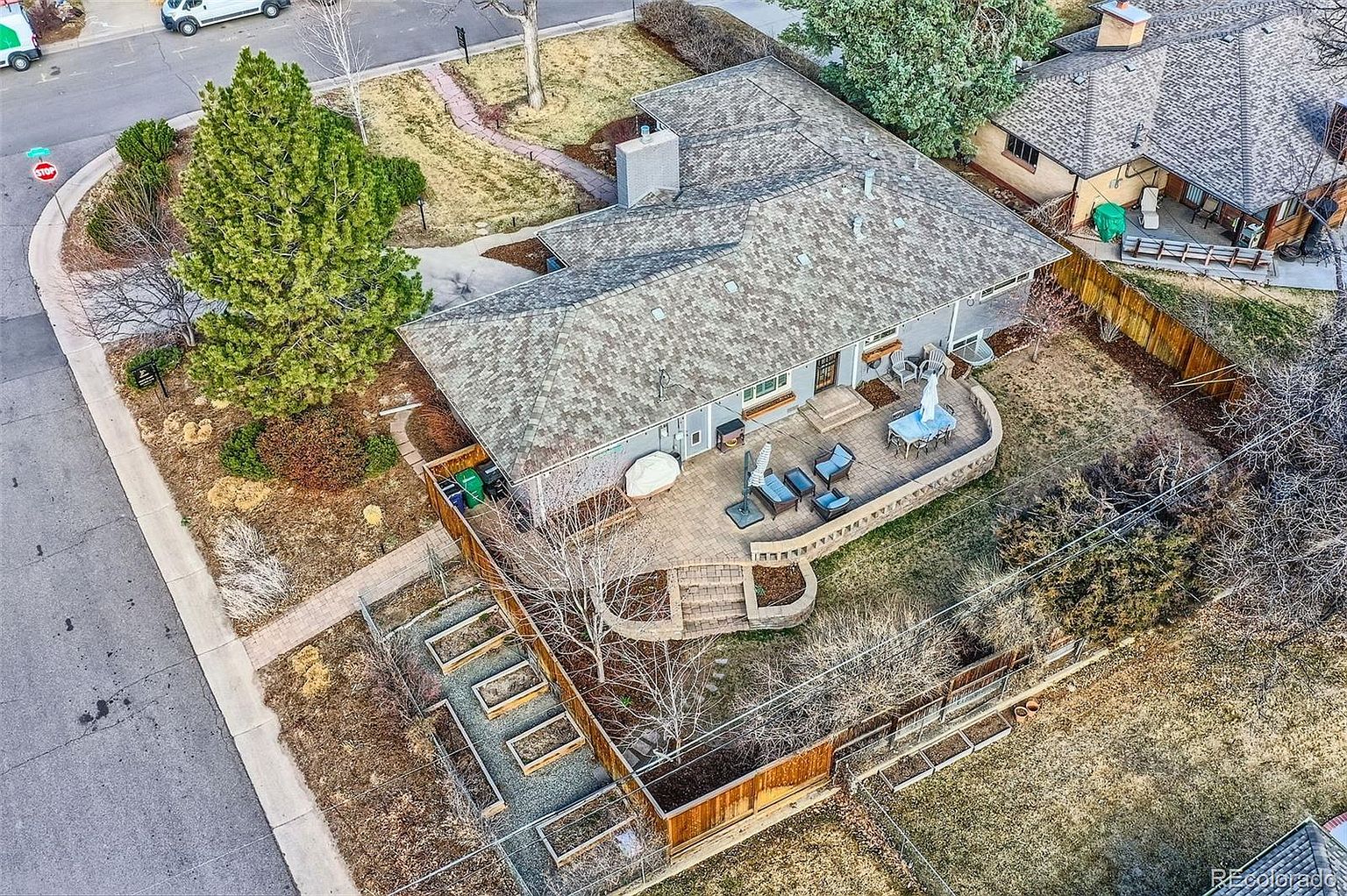 This aerial shot showcases a well-maintained single-family home with a spacious backyard featuring a patio area with outdoor furniture, a garden, and mature trees. The house has a gray roof and light-colored siding, complemented by a brick patio and a fenced yard. The overall impression is one of a comfortable and inviting property with ample outdoor living space.