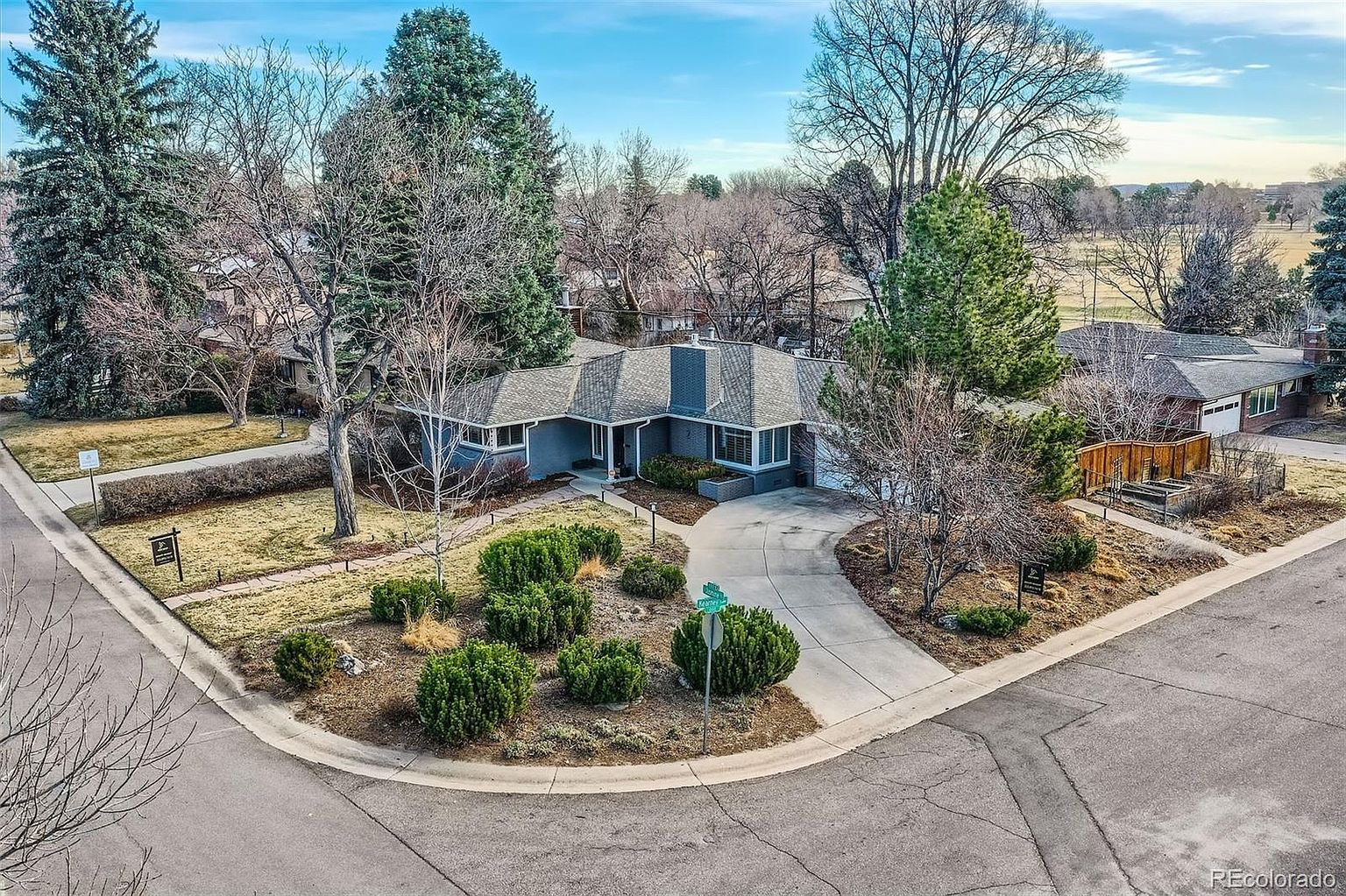 This aerial view showcases a well-maintained single-story home with a gray exterior and a neatly landscaped front yard. A curved driveway leads to the attached garage, and mature trees surround the property, providing shade and privacy. The overall impression is one of a charming and established residence in a desirable neighborhood.