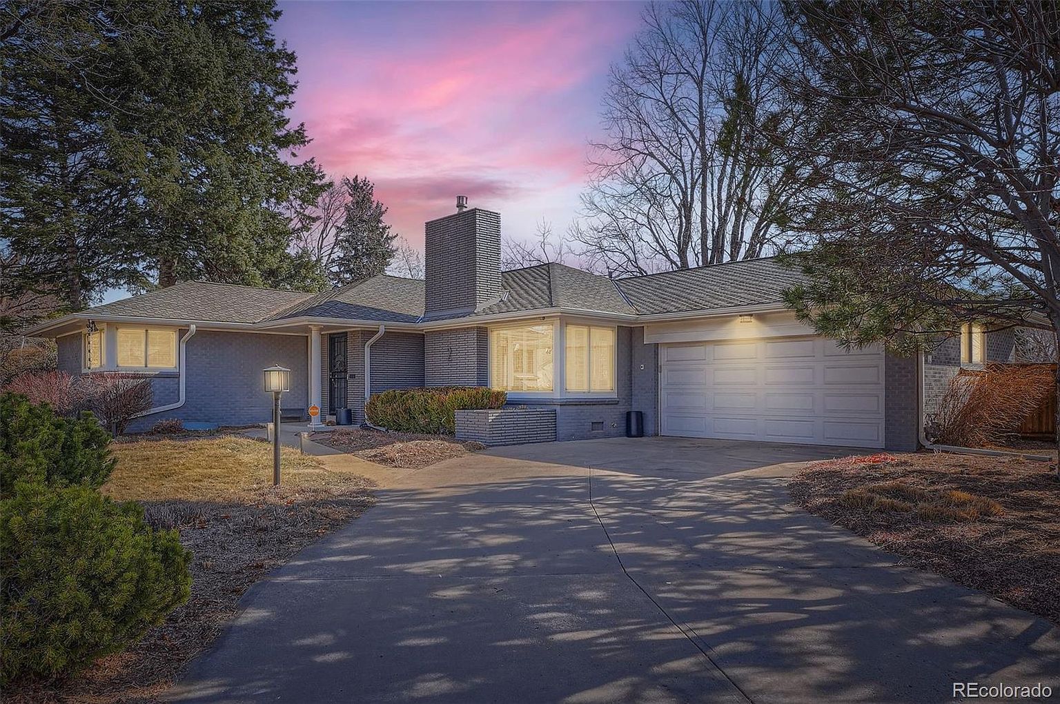 The image showcases the front exterior of a single-story brick house with a well-maintained lawn and mature trees. A concrete driveway leads to an attached two-car garage. The house features a prominent chimney and large windows, suggesting ample natural light inside, and the overall impression is one of a comfortable and established family home.
