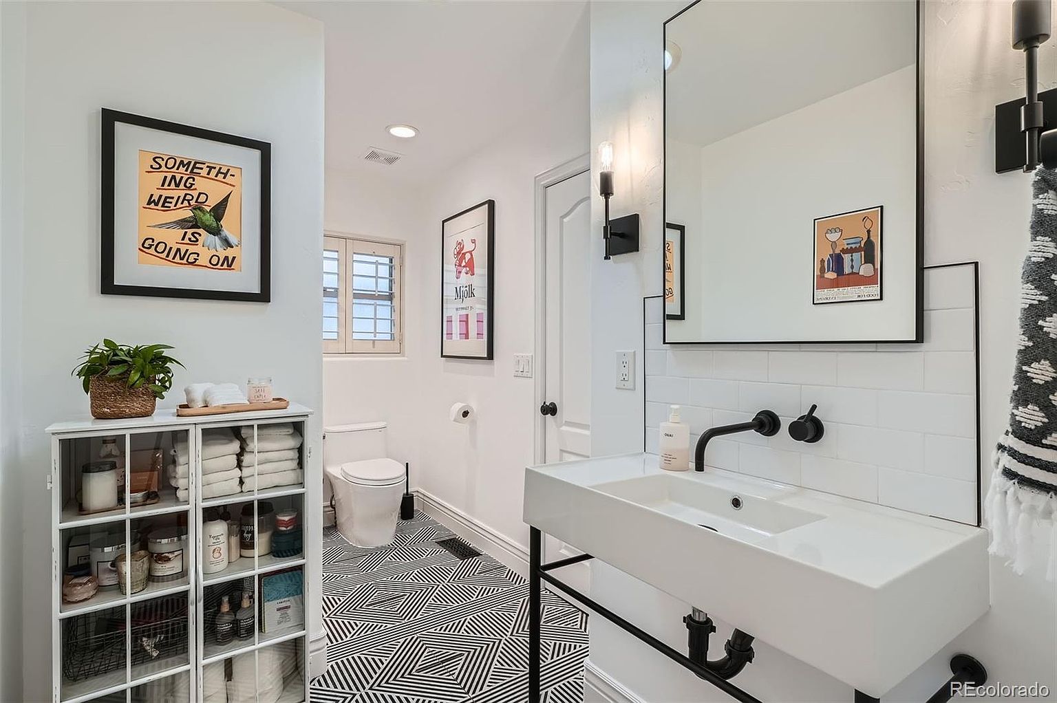 This is a stylish guest bathroom featuring a modern floating sink with black fixtures, complemented by a large mirror and subway tile backsplash. The black and white geometric floor tiles add a contemporary touch, while framed artwork and a storage cabinet provide visual interest and functionality. The overall impression is clean, modern, and well-designed.