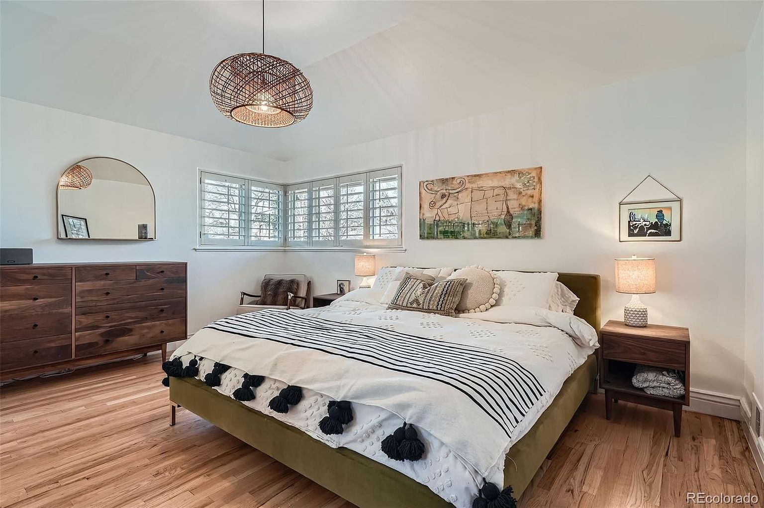 This is a well-lit primary bedroom featuring a modern aesthetic with hardwood floors and white walls. The room includes a large bed with a striped blanket and decorative pillows, a wooden dresser with a mirror, and a nightstand with a lamp. A woven pendant light hangs from the ceiling, adding a touch of texture to the space.