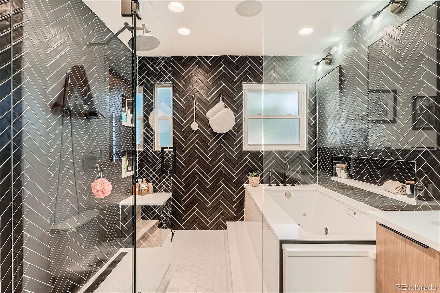 This is a modern primary bathroom featuring a walk-in shower with glass enclosure and a separate soaking tub. The walls are adorned with dark herringbone tile, complemented by white hexagonal floor tiles. The bathroom also includes a built-in shelf and a window providing natural light, creating a luxurious and spa-like atmosphere.