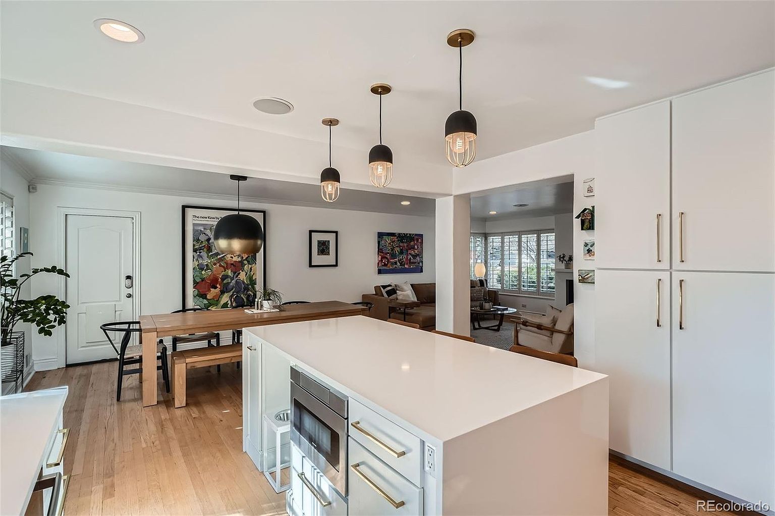 This is an interior shot of a modern kitchen with hardwood floors and white walls. The kitchen features a large white island with gold hardware, stainless steel appliances, and pendant lighting. The space opens to a dining area with a wooden table and a living room with comfortable seating, creating an inviting and open atmosphere.