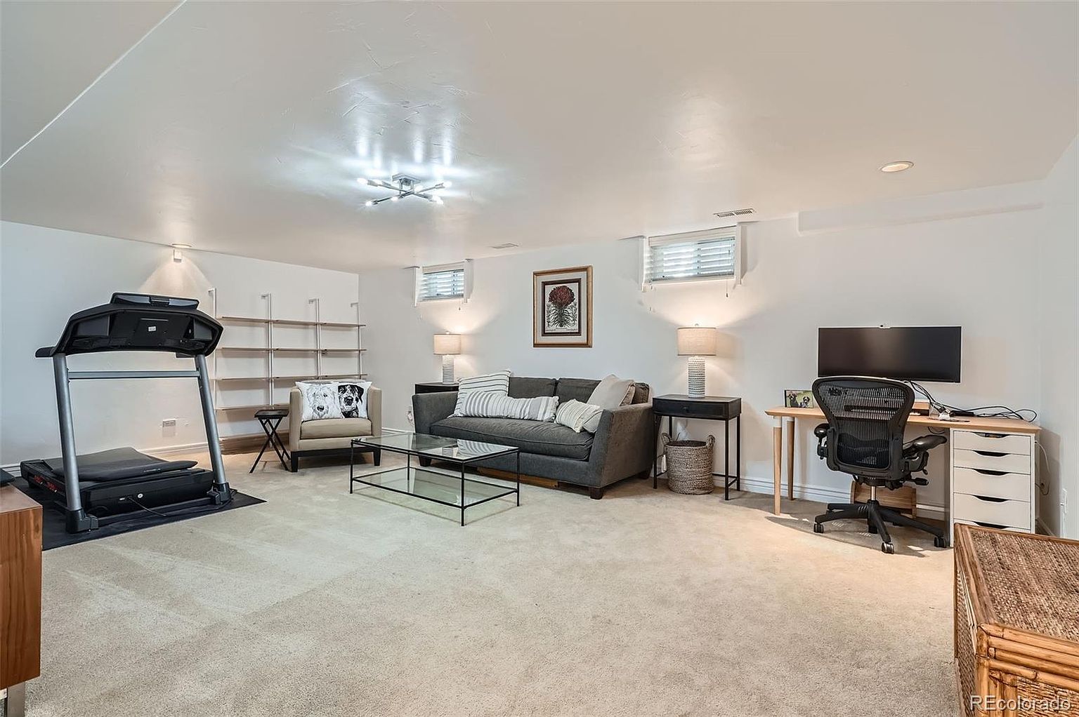 This is a well-lit basement area designed for both relaxation and functionality. The space includes a gray sofa with decorative pillows, a glass coffee table, a desk setup with a computer and office chair, and a treadmill for exercise. The room is carpeted and features white walls, creating a clean and versatile environment.