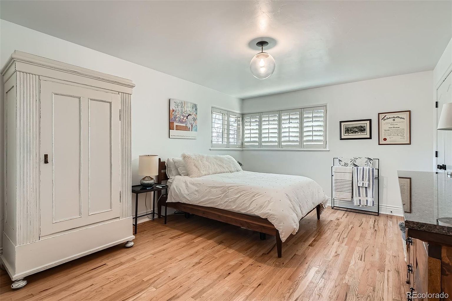This is a bright and airy primary bedroom featuring hardwood floors, white walls, and natural light streaming through shuttered windows. A large white wardrobe stands against one wall, while a wooden-framed bed with white bedding and fluffy pillows anchors the room. Artwork and framed documents adorn the walls, adding a personal touch to the space.