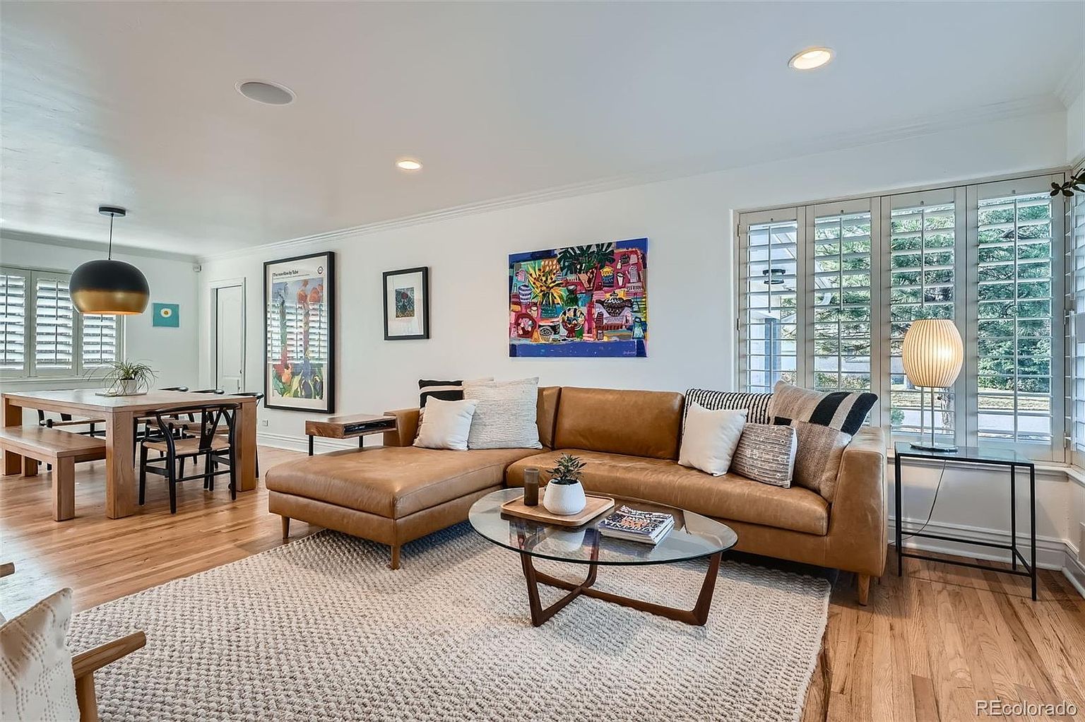This is an interior shot of a living room featuring a brown leather sectional sofa, a glass-topped coffee table, and a light-colored area rug. The room is well-lit with natural light coming through shuttered windows, and the walls are adorned with colorful artwork. The overall impression is a comfortable and stylish living space.
