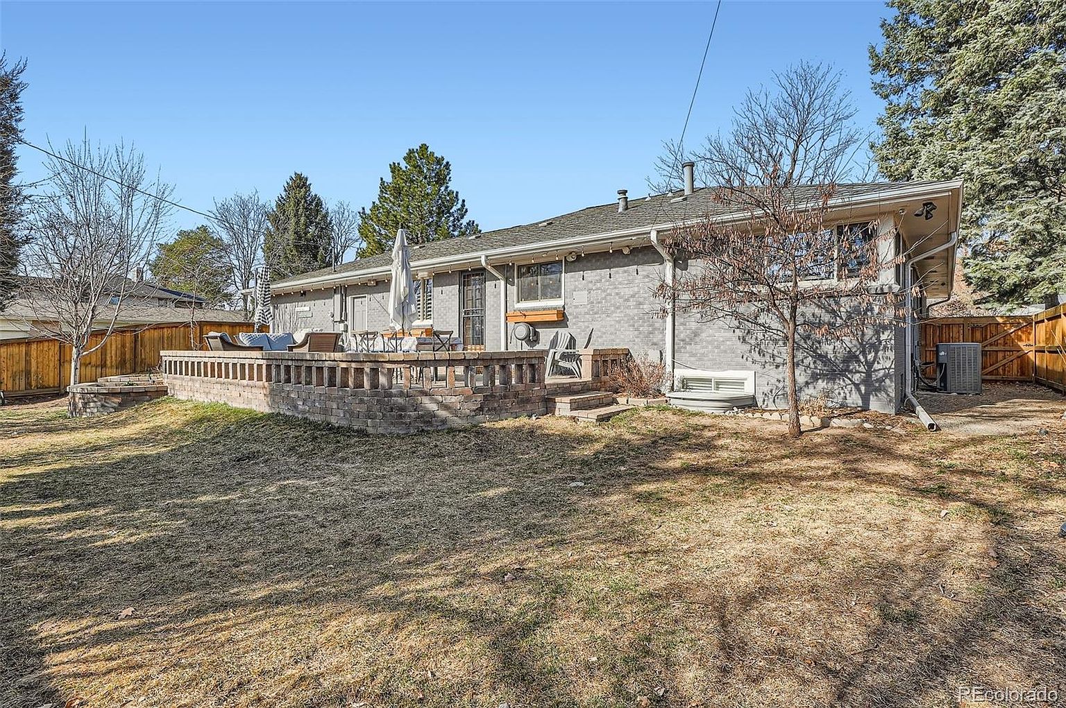 This image showcases the rear exterior of a single-story home, featuring a brick patio with outdoor furniture, a well-maintained lawn, and mature trees. The house has a gray brick facade and a low-pitched roof. The backyard is enclosed by a wooden fence, providing privacy and a sense of seclusion.
