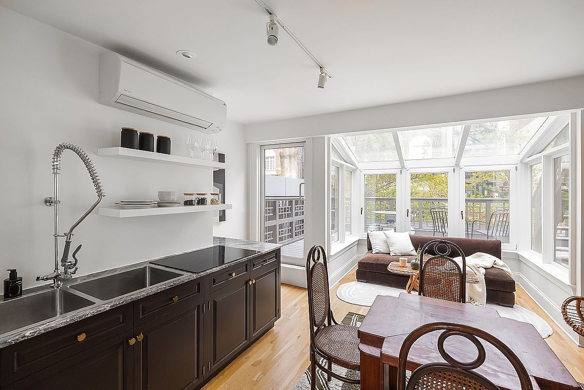 This interior shot showcases a combined kitchen and dining area, featuring dark wood cabinetry with a marble countertop and stainless steel sink. The kitchen area transitions into a bright sunroom-style dining space with a brown sofa and wicker chairs, offering a cozy and inviting atmosphere. The room is well-lit with natural light streaming through the surrounding windows.