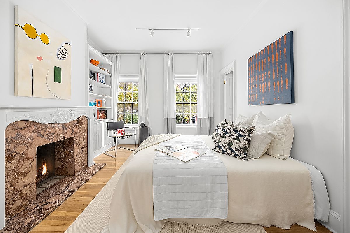 This is a bright and cozy bedroom featuring a daybed with decorative pillows and a throw, complemented by a marble fireplace and built-in bookshelves. Natural light streams in through the windows, enhancing the room's inviting atmosphere. Artwork adorns the walls, adding a touch of personality to the space.