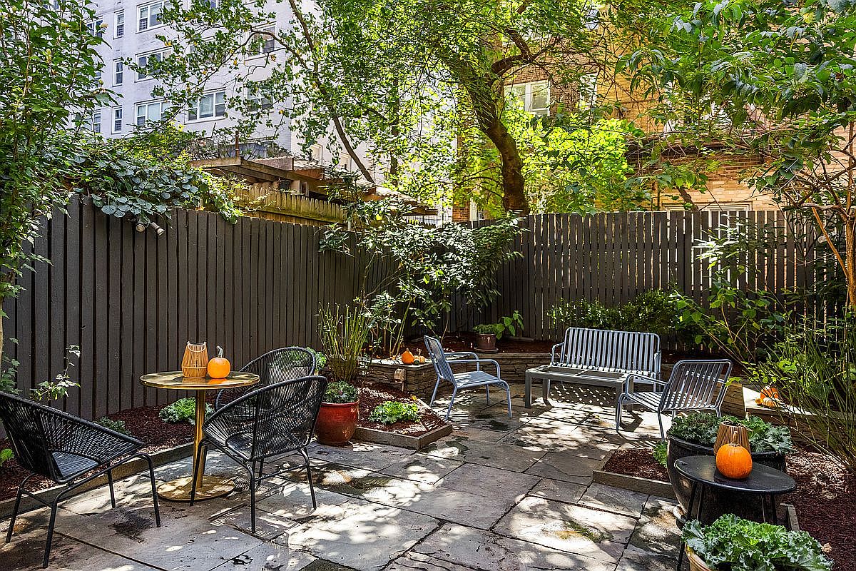 This is a charming backyard garden featuring a stone patio with outdoor seating, including chairs and a bench. The space is enclosed by a dark wooden fence and lush greenery, creating a private and tranquil atmosphere. Decorative pumpkins add a touch of seasonal charm.