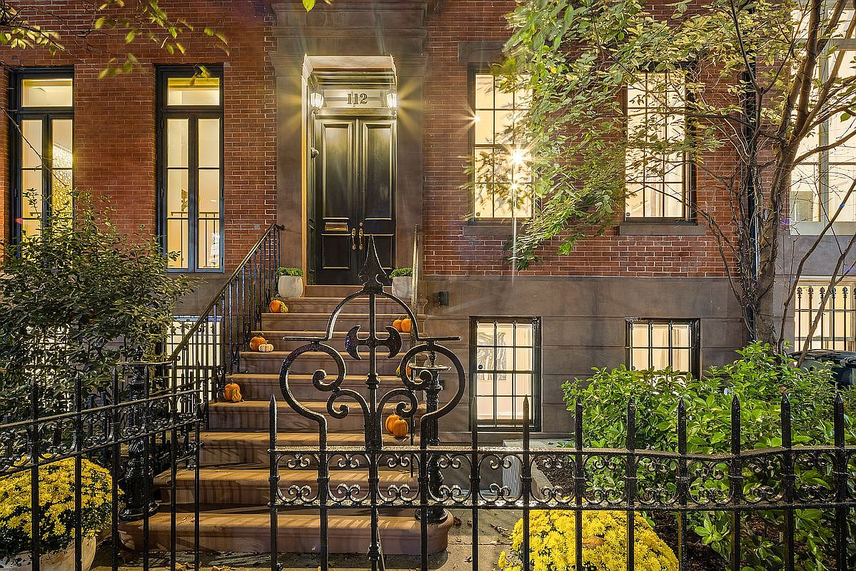 This is an inviting exterior shot of a brick townhouse, focusing on the entryway. The entrance features a black door with the number '112' above it, flanked by sconce lighting. Decorative wrought iron railings lead up the steps, which are adorned with pumpkins, suggesting a fall theme. Yellow flowers in pots add a pop of color to the foreground, enhancing the curb appeal.