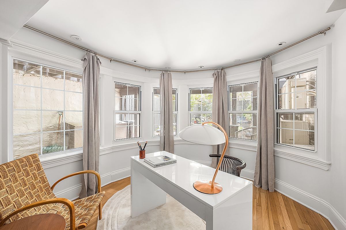 This is an interior shot of a bright and airy office space. The room features a curved wall of windows dressed with neutral-toned curtains, a modern white desk with a stylish lamp, and a woven chair. The hardwood floors and light-colored walls contribute to a clean and inviting atmosphere, perfect for a home office.