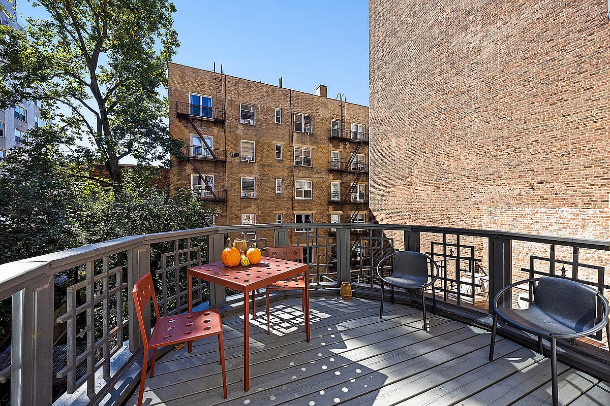 This image showcases a charming balcony setup, featuring a small orange table adorned with pumpkins, accompanied by matching chairs. The balcony has a wooden deck and a decorative railing, offering a cozy outdoor space. The backdrop includes a brick building and lush greenery, creating a pleasant urban oasis.