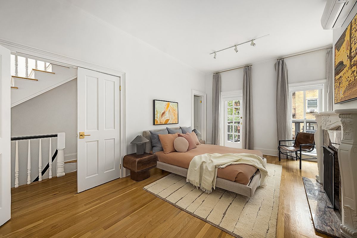 This primary bedroom features hardwood floors, white walls, and a neutral-toned bed with decorative pillows. Natural light streams in through a window with gray curtains, and a fireplace adds a touch of elegance. A doorway leads to a staircase, enhancing the room's spacious feel.