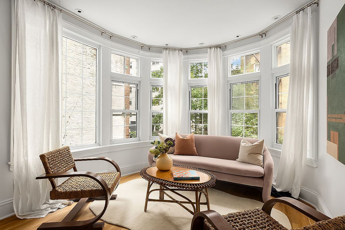 This is an interior shot of a living room featuring a curved bay window with sheer white curtains. The room is furnished with a light pink sofa, two woven chairs, and a round coffee table on a light-colored rug. The overall impression is bright, airy, and inviting, with a focus on natural light and comfortable seating.