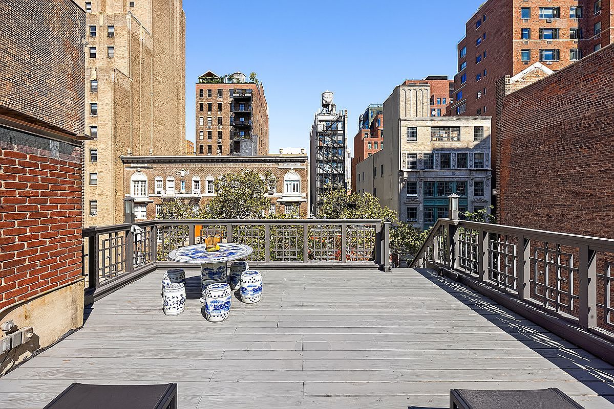 This image showcases a spacious rooftop deck with a gray wooden floor and a decorative railing. A round table with blue and white accents is surrounded by matching stools, creating a cozy outdoor seating area. The deck offers views of the surrounding cityscape, including brick buildings and a water tower, adding to the urban charm.