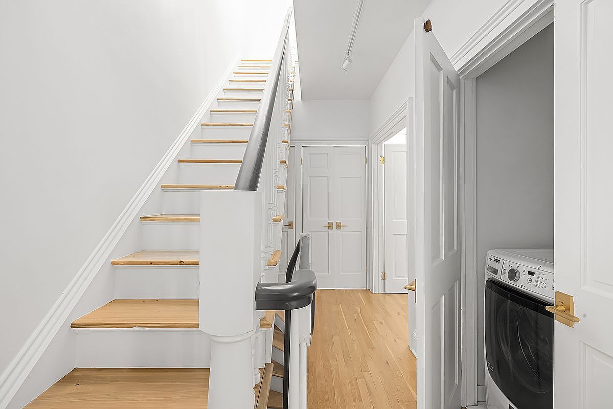 This interior shot showcases a well-lit hallway with a staircase leading upwards. The stairs feature wooden treads and white risers, complemented by a white banister with a dark handrail. A laundry area is visible through an open door, adding a practical element to the space, while the hardwood flooring and white walls create a clean and bright atmosphere.