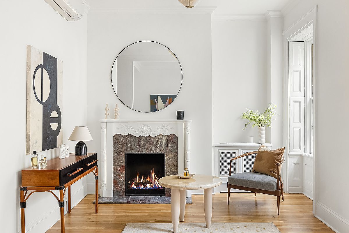 This is an interior shot of a living room featuring a fireplace with a round mirror above it. The room is decorated in a minimalist style with white walls, hardwood floors, and a mix of modern and vintage furniture. There is a console table with a lamp and artwork, a round coffee table, and a chair with a pillow, creating a cozy and inviting atmosphere.