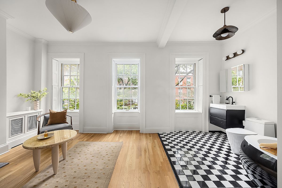 This is a bright and airy primary bathroom featuring a blend of classic and modern design elements. The room is well-lit by three large windows, and the flooring transitions from hardwood to a striking black and white checkered pattern in the bathing area. The bathroom includes a modern vanity, toilet, and a luxurious soaking tub, creating a spa-like atmosphere.