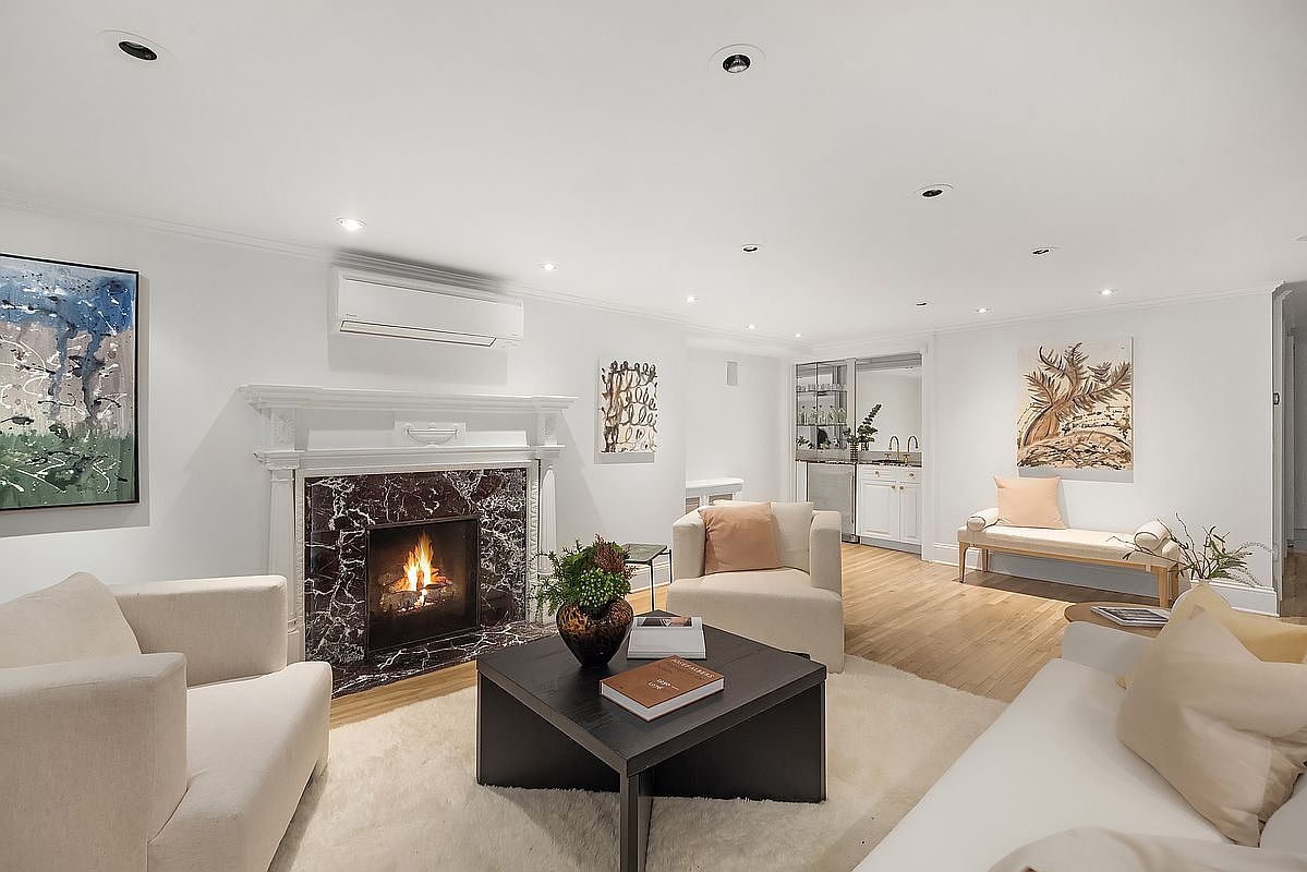 This is an interior shot of a living room featuring a fireplace with a marble surround, a dark wood coffee table, and neutral-toned seating. Artwork adorns the walls, and the room is well-lit with recessed lighting, creating a warm and inviting atmosphere. The perspective is from a medium distance, capturing the entire living space.