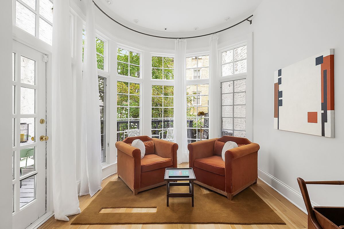 This is an interior shot of a living room featuring two rust-colored armchairs facing each other with a small table between them. The room has a curved wall of windows with sheer white curtains, allowing natural light to fill the space. A modern art piece hangs on the wall, and a brown rug covers the hardwood floor, creating a cozy and inviting atmosphere.