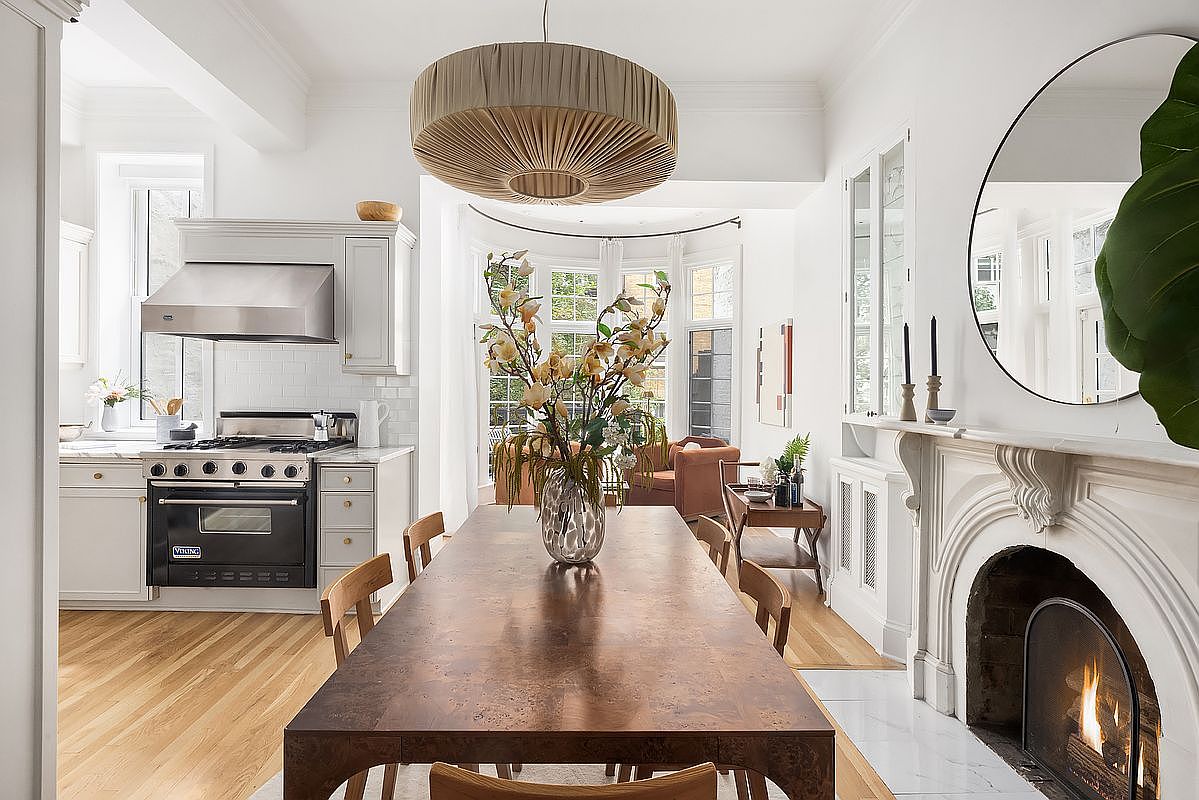 This interior shot showcases a dining room that seamlessly blends into the kitchen and living spaces. The focal point is a large wooden dining table adorned with a floral arrangement, illuminated by a unique pendant light. The room features a fireplace with a decorative mantel and a large round mirror, adding a touch of elegance and warmth to the space.