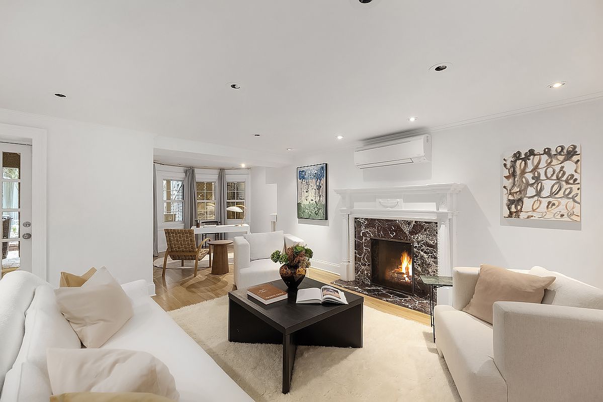 This is an interior shot of a well-lit living room featuring a fireplace with a marble surround, white sofas with beige pillows, and a dark wood coffee table with books and flowers. The room has white walls, recessed lighting, and artwork, creating a bright and inviting atmosphere. A bay window area with a desk and chair is visible in the background, adding to the room's functionality and charm.