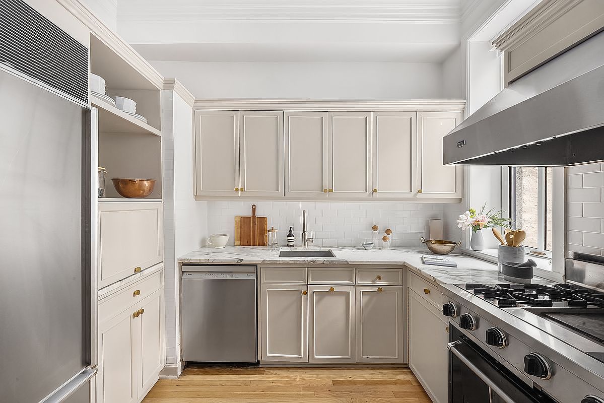This is a well-lit kitchen featuring custom cabinetry in a neutral tone with gold hardware. Stainless steel appliances, including a refrigerator, dishwasher, and range, are present. The countertops are marble, and the backsplash is white subway tile, creating a clean and classic look.
