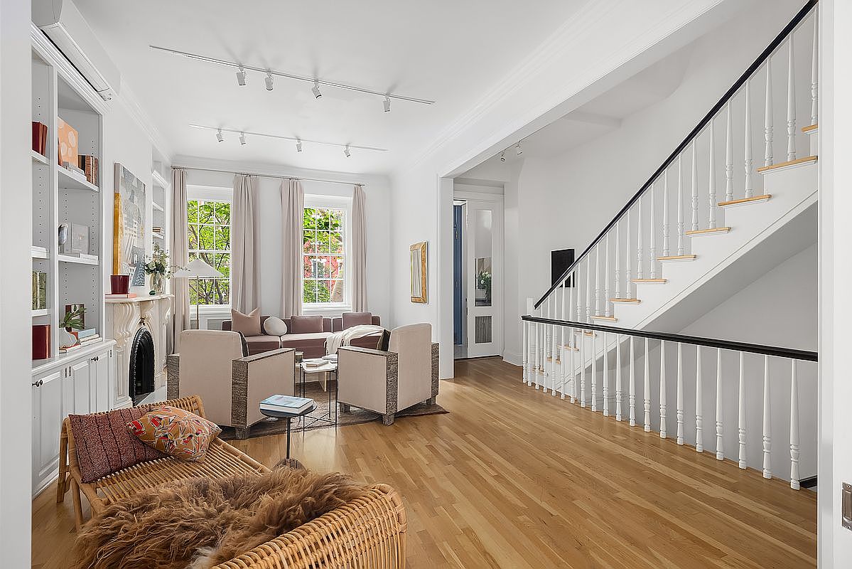 This is a bright and airy living room featuring hardwood floors, white walls, and a staircase with black handrails. The room is furnished with a sofa, two armchairs, and a wicker chaise lounge. Natural light floods the space through large windows, creating a welcoming and elegant atmosphere.