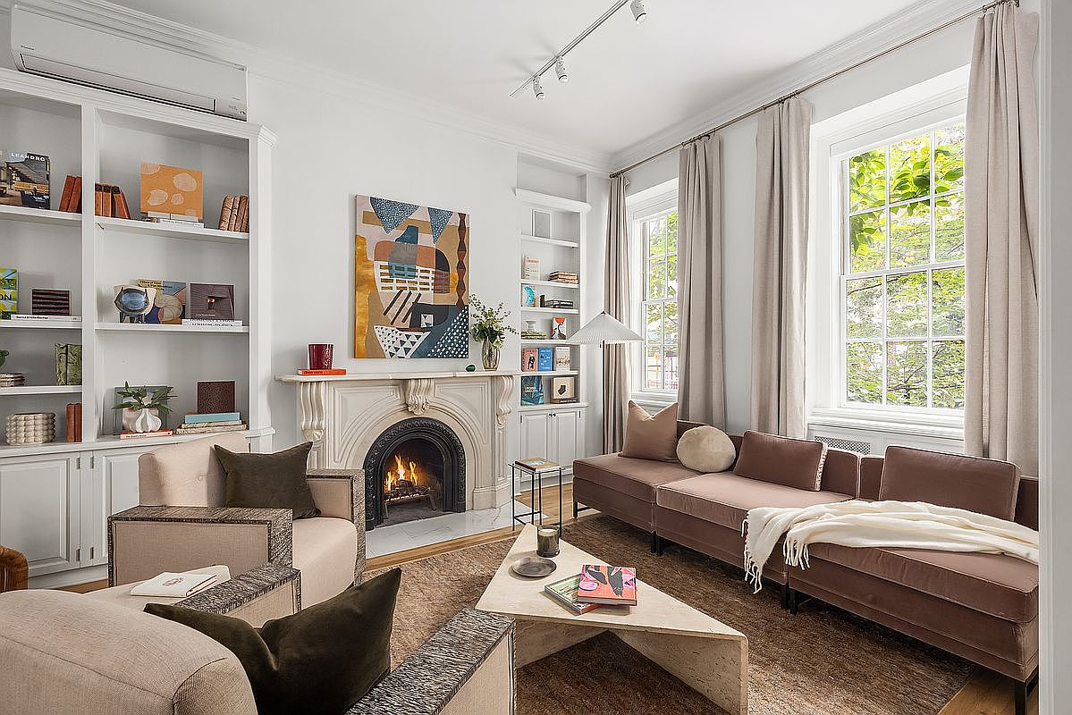 This is an inviting living room featuring a fireplace with a decorative mantel, flanked by built-in bookshelves. The room is furnished with comfortable seating, including armchairs and a sectional sofa, arranged around a modern coffee table. Natural light streams in through large windows, complemented by neutral-toned curtains, creating a warm and elegant atmosphere.