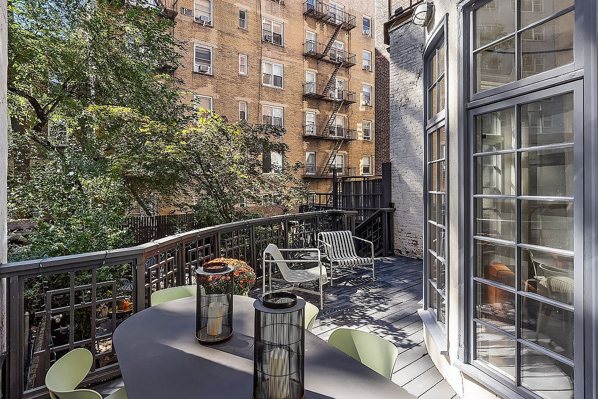 This image showcases a charming outdoor balcony or deck area, furnished with a modern dining table, chairs, and decorative lanterns. The space offers a view of a brick building with fire escapes and lush greenery, creating a cozy urban retreat. The deck's design and furnishings suggest a comfortable and stylish outdoor living space.