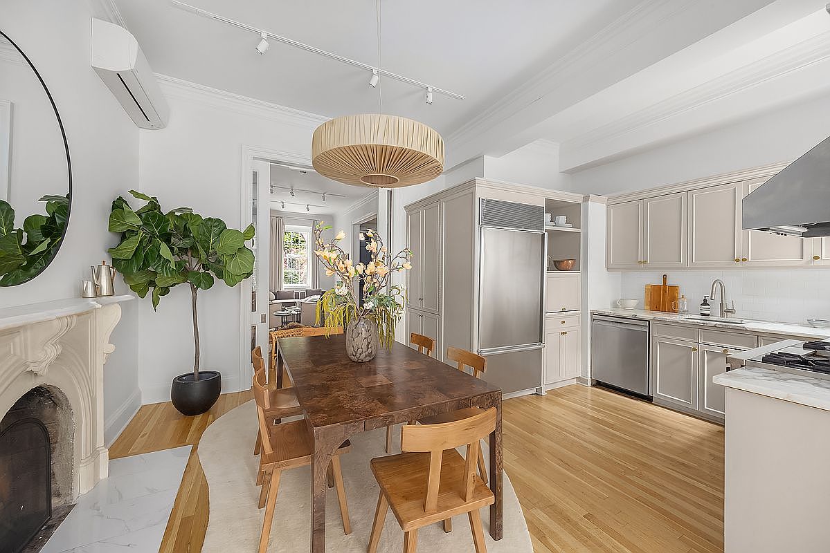 This interior shot showcases a well-lit kitchen and dining area, featuring a dark wood dining table surrounded by wooden chairs, set upon a light-colored rug. The kitchen area includes stainless steel appliances, light-colored cabinetry, and a marble countertop, while a decorative fireplace and a large potted plant add character to the space. The overall impression is one of a stylish and inviting home.